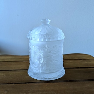 May include: A frosted glass jar with a lid, featuring embossed angel and star designs. The jar has a decorative base and a rounded top. The jar is sitting on a wooden surface.