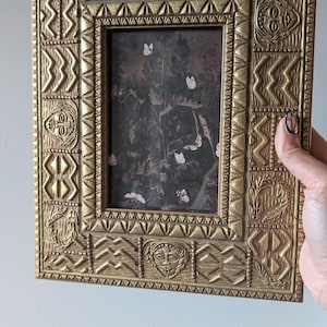 May include: Ornate gold-colored picture frame with a detailed, textured design. The frame surrounds a dark image with white butterflies. The frame has carved faces and geometric patterns.