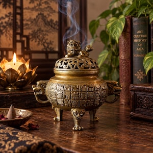 Peut inclure: Un brûle-encens en bronze métallique doté d'un corps sphérique orné de caractères chinois en relief, de deux anses latérales en forme de têtes d'animaux et de trois pieds. Le couvercle est surmonté d'une figurine de lion sculptée et dégage de la fumée.