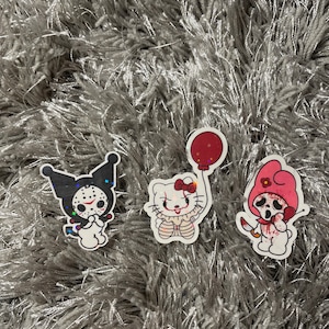 May include: Three vinyl stickers featuring a black and white Hello Kitty character with a bloody mask, a white Hello Kitty character with a red clown nose and a red balloon, and a pink Hello Kitty character with a bloody mask and a knife.