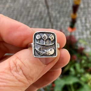 Könnte beinhalten: Ein Silberring mit rechteckiger Oberfläche mit detailliertem Design. Das Design umfasst drei stilisierte Bäume über einer Linie und einen Fisch darunter. Der Ring hat eine strukturierte, antike Oberfläche.