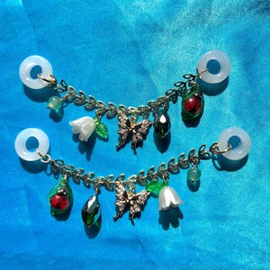 May include: Two pairs of white, circular earrings with gold-coloured chains. Each chain features a butterfly charm, a ladybird charm, a green gem, a white flower, and green leaves. The earrings are set against a blue background.