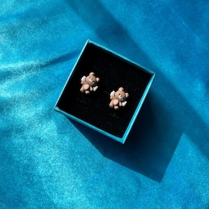 May include: Two small brown bear-shaped earrings with angel wings, displayed in a blue box on a blue background.