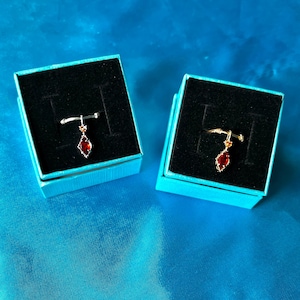 May include: Two ring boxes with a single ring in each. The ring on the left is silver with a red gemstone. The ring on the right is gold with a red gemstone.