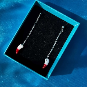 May include: A pair of silver dangle earrings with red and white enamel tooth charms. The earrings are displayed in a blue box with a black interior.