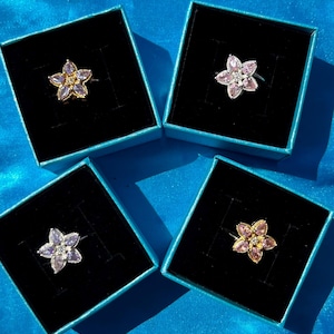 Può includere: Quattro anelli a forma di fiore in scatole nere individuali con bordi blu. Gli anelli presentano petali nei toni del rosa, viola e oro, con una gemma centrale. Le scatole sono disposte su una superficie blu.