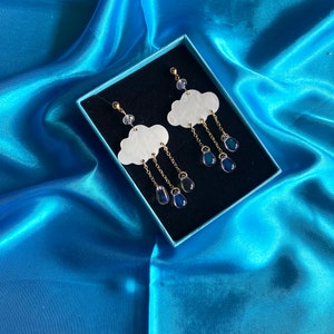 Puede incluir: Un par de pendientes en forma de nube blanca con cadenas doradas y abalorios en forma de lágrima azul iridiscente. Los pendientes se muestran en una caja de terciopelo azul.