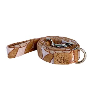 Puede incluir: Una correa para perro de color marrón y rosa con un clip plateado y una etiqueta de corcho que dice "The Little Daisy".
