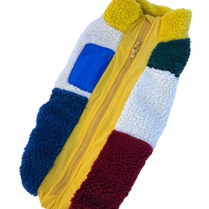 Op de afbeelding: Een veelkleurige fleecejas met een gele rits en hoge kraag. De jas heeft panelen in blauw, wit, geel, groen en bordeaux. Een blauwe zak is zichtbaar aan de linkerkant. De jas is gemaakt van een zacht, getextureerd materiaal.
