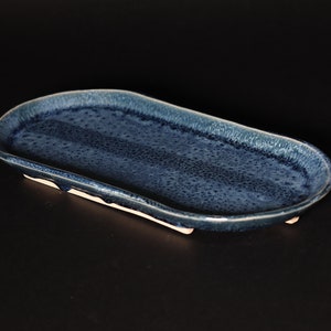 May include: A blue ceramic oval serving tray with a textured surface and a slightly raised rim. The tray has four small feet on the bottom.