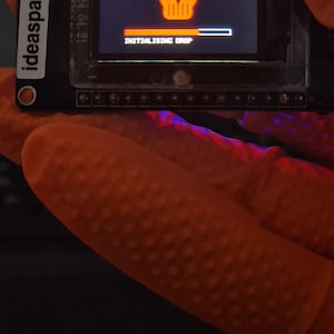 Op de afbeelding: Een zwarte Ideaspark elektronische module met een rechthoekig scherm dat een oranje schedelpictogram, een voortgangsbalk en de tekst INITIALISING DROP weergeeft. De datum 2024.04.21 staat verticaal gedrukt op de linkerzijde van het frame.