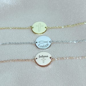 Könnte beinhalten: Drei silberne ovale Charm-Armbänder mit verschiedenen Gravuren. Das obere Armband hat ein Kreuz, das mittlere Armband ein Fischsymbol und den Namen "Luisa" und das untere Armband ein Kreuz und den Namen "Juliana".