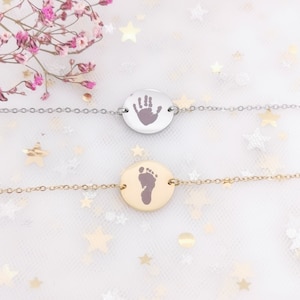 Könnte beinhalten: Zwei silberne und goldene Halsketten mit runden Anhängern, die einen Handabdruck und einen Fußabdruck zeigen. Die Halsketten liegen auf einem weißen Hintergrund mit kleinen silbernen Sternen.