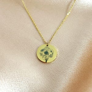 Peut inclure: Un collier dor&eacute; dot&eacute; d'un pendentif circulaire grav&eacute; d'une illustration botanique d&eacute;licate aux lignes sombres repr&eacute;sentant un bouquet de fleurs. Le pendentif est suspendu &agrave; une fine cha&icirc;ne dor&eacute;e assortie compos&eacute;e de petits maillons uniformes.