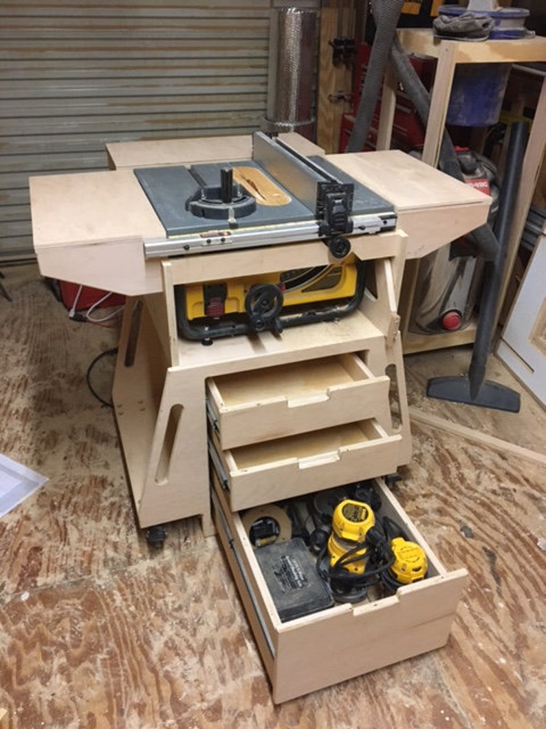 May include: A wooden table saw stand with three drawers. The top drawer is open and contains power tools, including a router and a sander. The table saw is on top of the stand and has a black top and a silver blade.