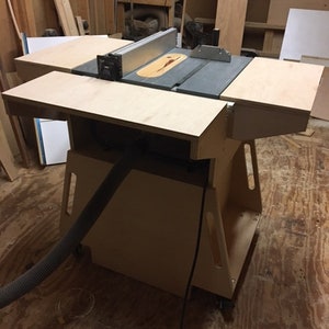 May include: A wooden table saw stand with a gray table saw on top. The stand has a black dust collection hose attached to it. The stand is on a wooden floor.
