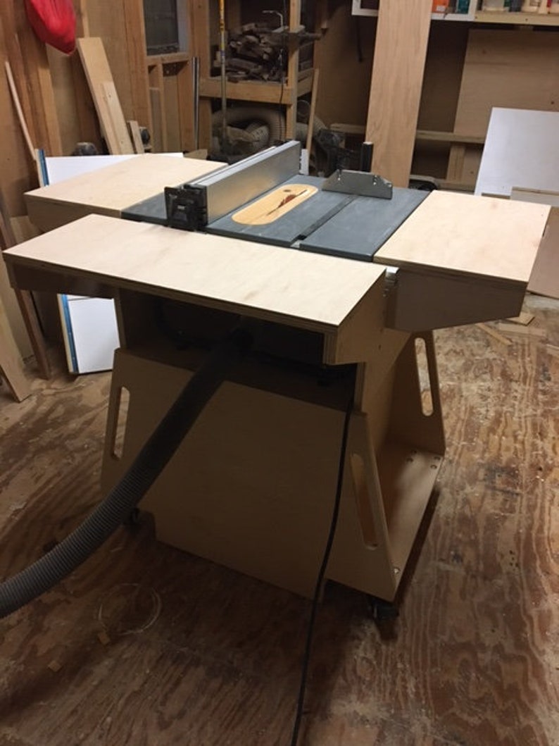 May include: A wooden table saw stand with a gray table saw on top. The stand has a black dust collection hose attached to it. The stand is on a wooden floor.
