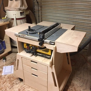 May include: A wooden table saw stand with three drawers and wheels. The table saw is mounted on top of the stand and has a black and silver blade.