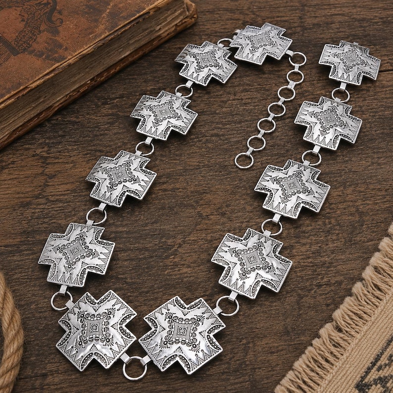 May include: A silver-toned metal chain belt featuring a series of repeating cross-shaped links. Each link is decorated with intricate engraved patterns and a central square motif. The belt includes a circular link adjustment chain at one end.