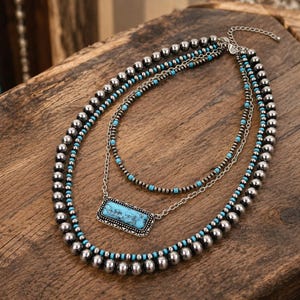 May include: A multi-strand necklace featuring large silver-toned metallic beads, smaller turquoise-colored beads, and a central rectangular pendant with a turquoise stone inlay set in a decorative silver-toned frame with a metal chain link.
