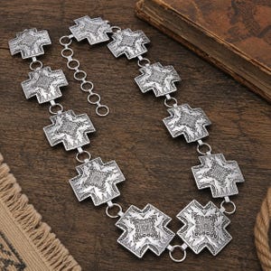 May include: A silver-toned metal chain belt featuring a series of repeating cross-shaped links. Each link displays an intricate, embossed geometric pattern. The belt includes a circular link adjustment chain at one end for sizing flexibility.