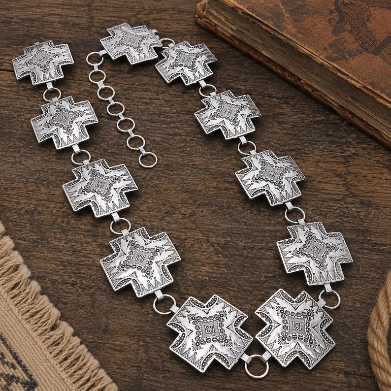 May include: A silver-toned metal chain belt featuring a series of repeating cross-shaped links. Each link displays an intricate, embossed geometric pattern. The belt includes a circular link adjustment chain at one end for sizing flexibility.