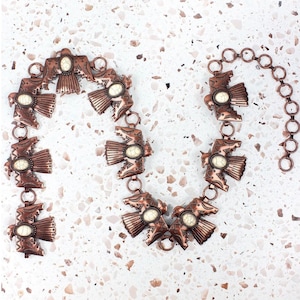 Pode incluir: Um cinto de tom cobre com uma corrente e pingentes em forma de águia. Cada pingente tem uma pedra branca no centro.