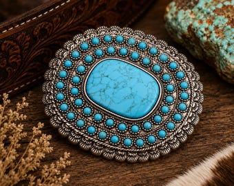 May include: An oval belt buckle featuring a large, central rectangular turquoise stone with dark veining, surrounded by two concentric rings of small, round turquoise cabochons set in an ornate, antiqued silver-tone metal frame with scalloped edges.