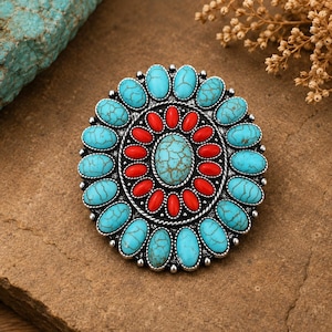 Puede incluir: Un broche circular que presenta una piedra turquesa ovalada central con vetas marrones, rodeada por un anillo de pequeñas piedras ovaladas rojas y un borde exterior de piedras turquesas ovaladas más grandes, montado en un marco de metal plateado.