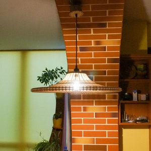 May include: A brown woven pendant light fixture hangs from the ceiling in a room with a brick wall and a window with blinds. The light fixture has a wide, flat shade.