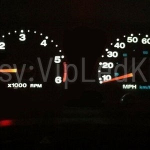 Może przedstawiać: Czarna deska rozdzielcza z białymi wskaźnikami, w tym prędkościomierzem, kt&oacute;ry wskazuje do 100 mph, i obrotomierzem, kt&oacute;ry wskazuje do 6 000 obr./min. Wskaźniki są podświetlone na biało i niebiesko.