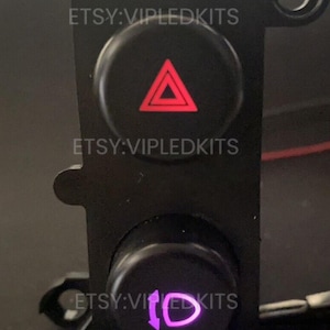 May include: Black automotive switch panel with two illuminated buttons. The top button features a red triangle, and the bottom button displays a purple symbol. The text "etsy:VIPLEDKITS" is visible. Automotive parts.