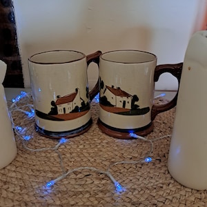 Puede incluir: Dos tazas de cerámica con base crema, detalles marrones y asa. Cada taza presenta una escena pintada de una casa con tejado marrón y árboles verdes. Las tazas están sobre una superficie tejida con luces de cadena azules.