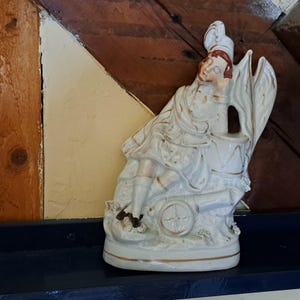 Op de afbeelding: Een wit keramiek beeldje van een persoon die op een kanon zit. Het beeldje is geschilderd met gouden accenten en heeft een witte hoed. Het beeldje staat op een plank.