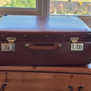 May include: A rectangular brown suitcase featuring dark brown reinforced corner caps, a central leather handle, and two silver-toned metal latches with integrated keyholes. The exterior surface shows a smooth finish with visible signs of wear.