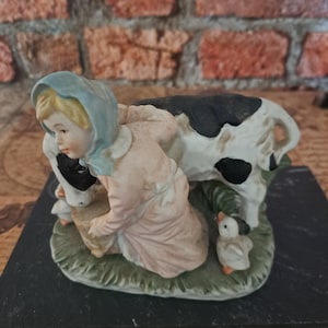 Può includere: Una statuetta in porcellana raffigurante una persona con una cuffia azzurra e abiti rosa inginocchiata accanto a una mucca bianca e nera. Due piccoli anatroccoli bianchi sono posizionati vicino alla persona su una base verde.
