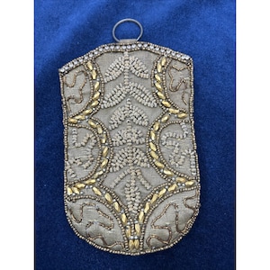 May include: A vintage, rectangular, beaded pouch with a silver metal ring at the top. The pouch features an intricate design of gold and silver beads, creating floral and geometric patterns on a light tan fabric. The edges are also adorned with gold beads.