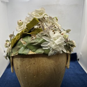 May include: A decorative hat featuring a cream-colored base adorned with white fabric flowers, green fabric leaves, and a prominent olive green velvet ribbon bow. The structure includes a delicate white lace overlay covering the top surface.