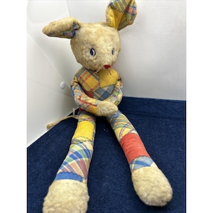 May include: A vintage stuffed bunny rabbit with long legs and ears. The bunny has a beige head and limbs, with blue eyes and a red nose. The body and legs are patchwork with yellow, red, and blue plaid fabric. A charming, nostalgic toy.