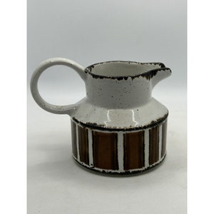 May include: A ceramic pitcher with a white speckled top and a brown and white striped base. The pitcher has a rounded handle and a spout for pouring. The design is a mix of modern and rustic styles.
