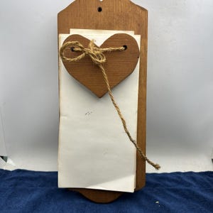 May include: Wooden wall-mounted memo board with a heart-shaped accent. The board is made of light brown wood and features a heart-shaped cutout with a twine bow. White paper is held in place by the heart and twine.