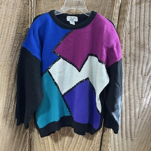 May include: A colorful sweater with a geometric design. The sweater features a black crew neck and sleeves, with a patchwork of blue, teal, purple, white, and magenta. Black sequins outline the geometric shapes. The label reads "Victoria Jones Woman."