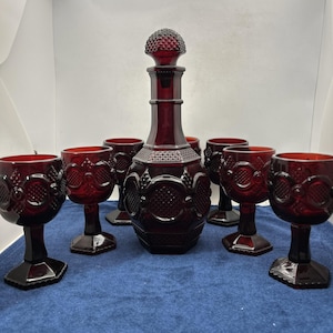 May include: A set of deep red glass drinkware consisting of a tall decanter with a matching stopper and six stemmed goblets. Each piece features a textured hobnail and geometric pattern across the surface, with a hexagonal base on the goblets.