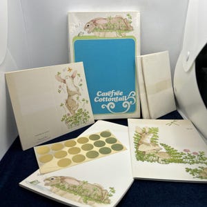 May include: A collection of vintage stationery featuring illustrations of rabbits and clovers. The set includes notepads, cards, and stickers. The notepad has a blue background with the words "Carefree Cottontail". The illustrations have a soft, pastel color palette.