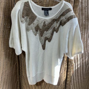 May include: Off-white short-sleeve sweater with decorative brown and tan accents. The accents are embellished with small, gold-colored studs. The sweater has a scoop neck and a relaxed fit.