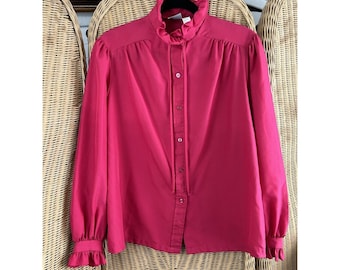 May include: A bright pink long-sleeved blouse with a ruffled collar and cuffs. The blouse has a button-down front and a tie detail at the neck. The fabric appears to be a lightweight material, and the blouse is hanging on a black hanger.