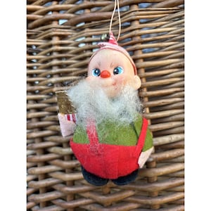 May include: A vintage Christmas ornament shaped like a gnome. The gnome has a white beard, rosy cheeks, and blue eyes. It wears a green shirt, red overalls, and a red and white striped hat. The ornament is suspended by a thin gold string.