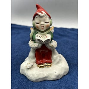 May include: A ceramic figurine of a singing elf in a red hat and pants, green scarf, and gold-trimmed shoes. The elf is holding a book and is standing on a snowy base with a small bird. The figurine has a vintage, Christmas theme.
