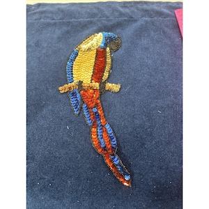 May include: A parrot-shaped patch crafted from sequins in vibrant shades of gold, red, and blue. The bird is depicted perched on a branch, with its long tail extending downward. The design features intricate beadwork outlining the bird's form.