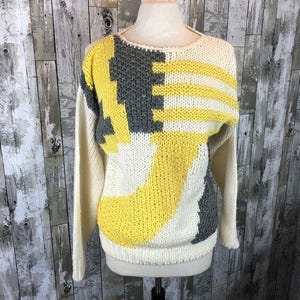 May include: Cream-colored sweater with a geometric design in yellow and gray. The sweater features a crew neck and long sleeves. The design includes horizontal stripes and angular shapes, creating a bold, retro look.